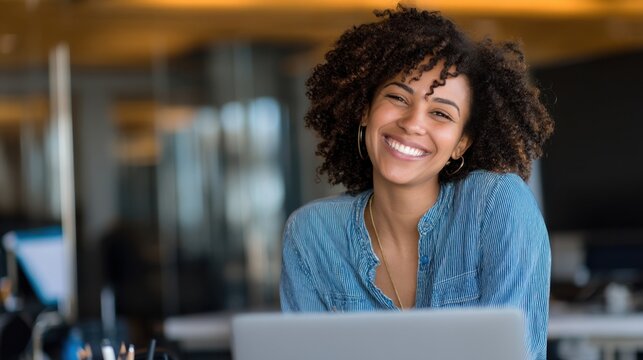 A woman sits at a desk with a laptop in a modern office. She smiles while working. The background shows a bright office space with large windows.