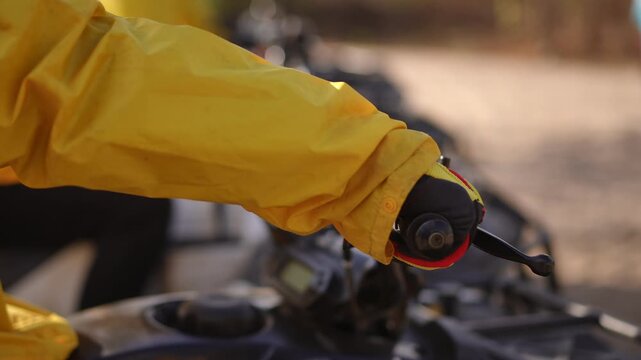Gloved hand on handlebar throttle preparing lineup of quads, yellow jacket sleeve and textured grip in sunlight, twisting motion signals imminent start, shallow
