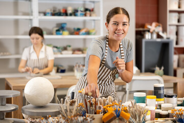 Young woman choosing brush to paint ceramic products in workshop