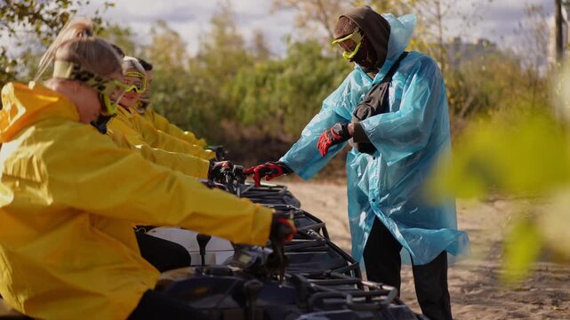 Quad riders in yellow jackets receive instruction from guide in blue poncho handing gloves next to lined ATVs on muddy trail, closeup of handoff, goggles