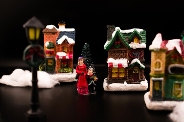 Cozy miniature Christmas village scene with caroling figures singing beside a snow-covered house and trees. Warm glowing windows, soft focus, and dark background create a nostalgic holiday atmosphere.