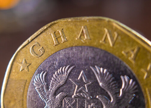 Close-up of the reverse of a bimetallic 2 Ghana Cedi coin on wood. The silver core shows the Coat of Arms with eagles and a star, while the golden outer ring is embossed with "GHANA" and stars.