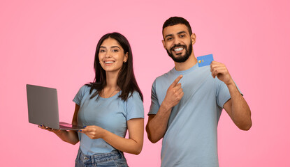 Two enthusiastic young adults stand together, one holding a laptop while the other displays a credit card.
