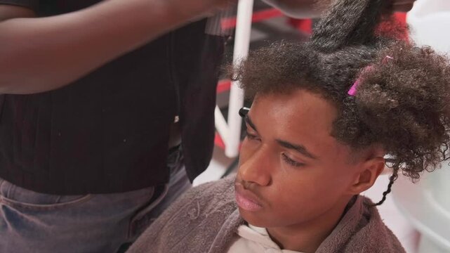 Teen boy at start of braiding process with afro hair sectioned, stylist marking and combing a strand with fine-tooth comb