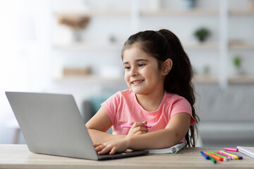 A cheerful girl sits at a table, using a laptop for online learning, holding a pen in her hand. Bright colors and a welcoming atmosphere create a fun study environment.