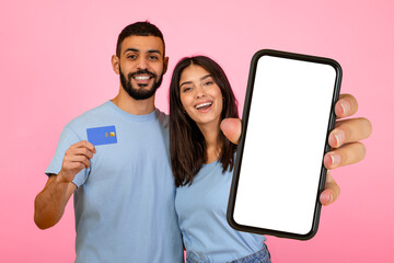 A young couple stands together, smiling brightly while holding a smartphone and a card. The background is a vibrant pink, adding a lively touch to their cheerful demeanor.