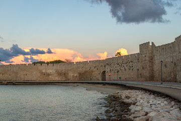 Sunset view of the medieval walls of Rhodes reflecting warm light over the sea. Historic fortifications along the coastline of Rhodes Island, Greece, with dramatic sky and calm water