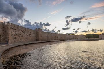 Sunset view of the medieval walls of Rhodes reflecting warm light over the sea. Historic fortifications along the coastline of Rhodes Island, Greece, with dramatic sky and calm water