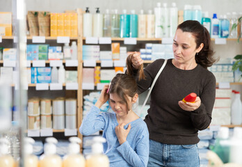 Woman stands in a pharmacy and checks her daughter's hair for lice. Child stands in the pharmacy with mother in search of remedies and shampoos for lice.