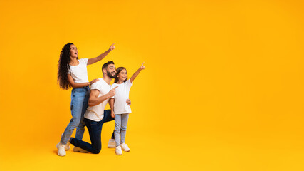 A joyful family stands against a vibrant yellow backdrop. They are playfully pointing at something out of view, sharing smiles and laughter. Parents and child are dressed casually. © Prostock-studio