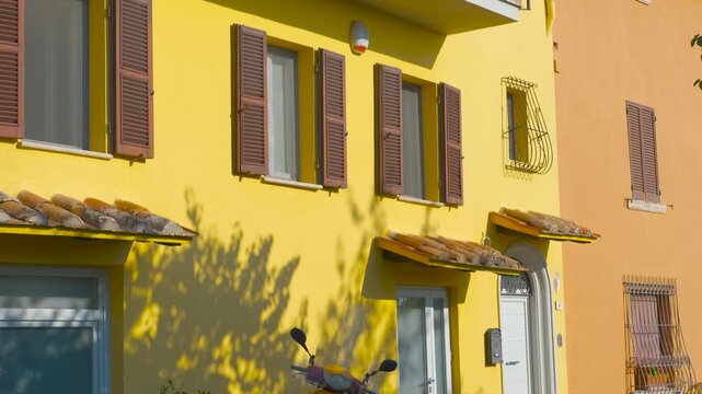 Yellow italian building facade revealing balconies. Facade of a traditional yellow italian apartment building with brown shutters, balconies with flowers, and a red scooter parked on the street