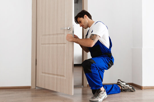 A young male locksmith in a blue uniform is kneeling while carefully installing a door knob on a wooden entrance door. The setting is a clean and modern interior, ideal for professional repairs.