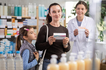 Interested woman with small daughter thoughtfully examining box of medicine while female pharmacist politely offering assistance in background in modern pharmacy