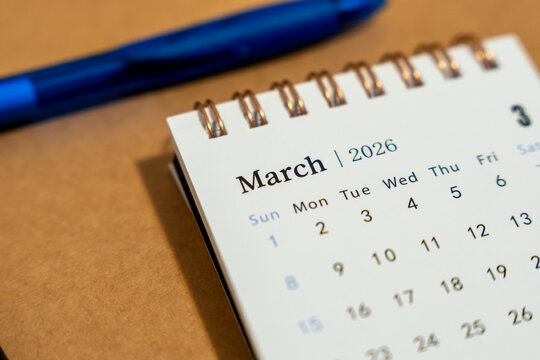Close-up view of a desk calendar displaying March 2026, along with a blue pen on a brown background. Ideal for organizing time management, planning, and office use.