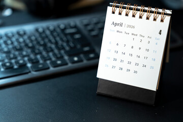 A desk calendar displaying April 2026 sits on a dark keyboard. The calendar highlights days of the week and numerical dates, suitable for organizational and planning purposes.