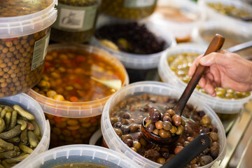 At vegetable shop, unrecognizable customer takes pickled olives out of bucket with skimmer spoon. Local farm and imported vegetables and fruits, self-service supermarket.