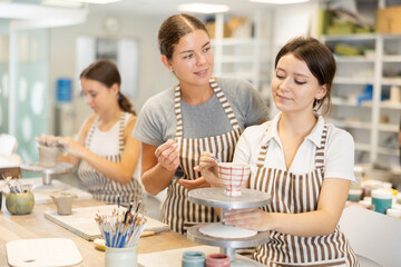 During master class, girl student paints clay vessel under guidance of female teacher. Mentor is next to student, makes comments and corrects inaccuracies