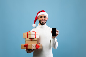 A cheerful man in a warm sweater wears a Santa hat as he showcases a smartphone in one hand while balancing several holiday gifts in the other. The background is a soft blue.