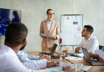 In a contemporary boardroom, a confident businesswoman presents ideas to her diverse team. Colleagues engage in discussion while reviewing strategies and analyzing data on laptops.