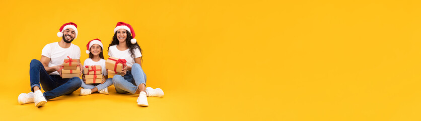 A joyful family of three sits on the floor wearing Santa hats. They hold wrapped gifts, surrounded by a festive atmosphere with a vibrant yellow backdrop.