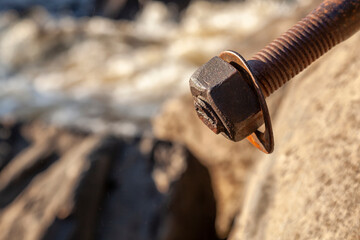 Macro view of an old rusty metal bolt with nut and washer fixed in rock, with a soft blurred river stream in the background