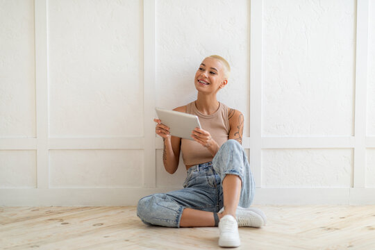 Smiling lady using digital tablet and looking aside at copy space, sitting on floor over white wall, free space. Happy woman posing with pad. Gadget lifestyle and online technology concept