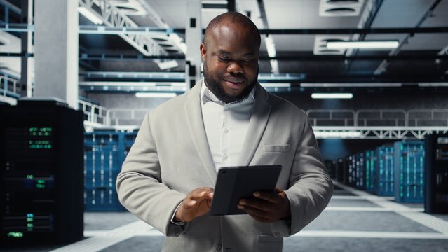 Smiling admin using tablet in in data center maintaining infrastructure for workloads. Joyful IT professional in server farm utilizing high tech server clusters for parallel processing, camera A
