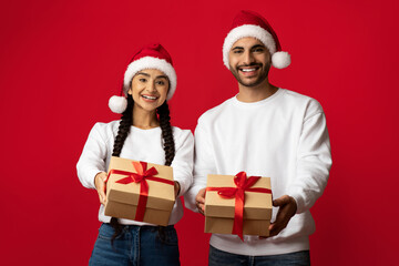 Joyful couple wears Santa hats and smiles widely while holding wrapped gifts. The festive scene features a vibrant red backdrop that enhances the holiday spirit.