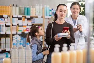 Interested woman in thirties using phone to scan barcode and examine ingredients of healing ointment for tween daughter, while professional pharmacist offering guidance at drugstore