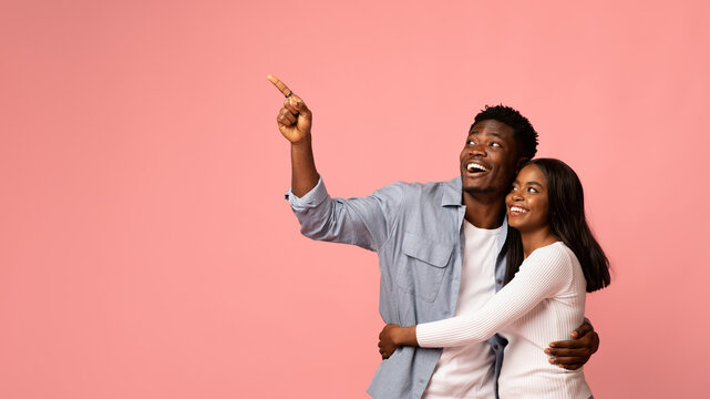 A joyful african american couple stands together, hugging and smiling while pointing at empty space. Their casual outfits and playful demeanor create a cheerful atmosphere against the pink backdrop. - Powered by Adobe
