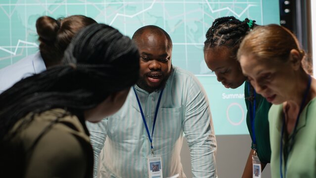 Diverse coworkers team participating in a late night briefing session, exchanging documents and reviewing finance updates. Aligning objectives through collaborative problem solving. Camera B.