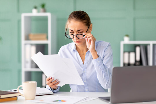 Female office worker sits at her desk focused on paperwork. She adjusts her eyeglasses, trying to read the documents clearly in her office. The environment is organized and professional.