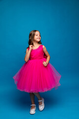 Naklejka premium Cute happy little girl in a pink fluffy princess dress has fun and dances on blue background. Kids Birthday party celebration concept. Happy Birthday, copy space. High quality photo