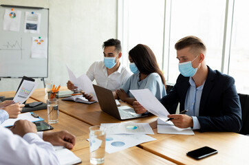 A diverse group of colleagues is working together in a bright office space. They are wearing face masks while discussing documents and strategies for company growth.