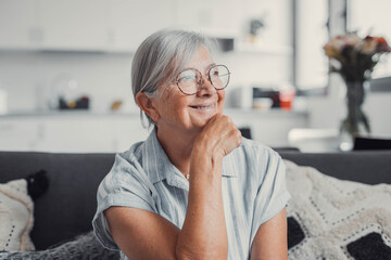 Elderly woman sitting on a sofa at home smiles as she looks out the window. She enjoys a peaceful...