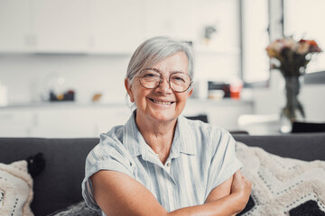 Happy elderly woman sits on a sofa at home and smiles at the camera during a video call. She feels connected and cheerful while enjoying a cozy and peaceful domestic setting.