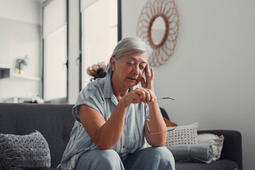 Elderly woman sits on a sofa at home suffering from a headache. She feels exhausted and stressed,...