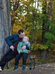 Fototapeta premium A father gently hugs and kisses his son while sitting on a swing during an outdoor walk with open copy space. Concept of Father's Day, tenderness, family love and emotional bonding.