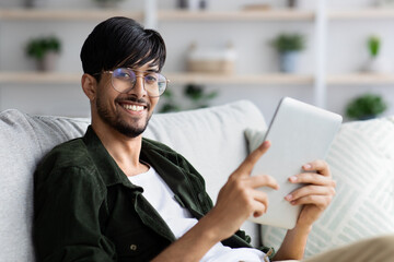 A young man sits comfortably on a couch, smiling while interacting with a tablet. The room is bright and decorated with plants, creating a warm atmosphere.