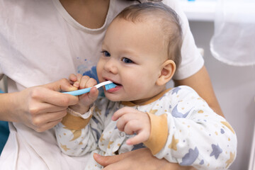 Happy cute one year old baby cleaning, brushing his first teeth with a blue toothbrush and toothpaste.  Young mother holding her child and taking care of personal hygiene. Prevention of dental disease