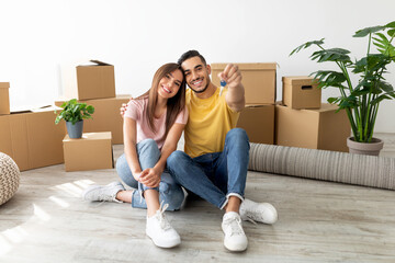 Happy couple sits on the floor of their new home, showing a set of keys. They are surrounded by moving boxes and indoor plants, sharing a joyful moment.