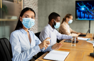 In a modern office, an Asian businesswoman applies disinfectant spray to her hands while her colleagues follow safety measures. The meeting proceeds under coronavirus precautions.