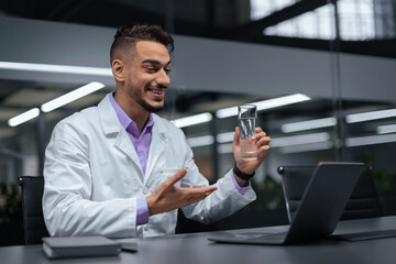 A doctor is smiling while talking to a patient over a video call. He holds a glass of water in one hand and is using the other hand to gesture. The setting is an office with modern decor.