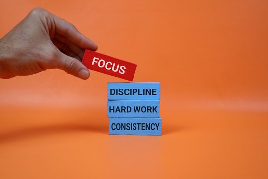 A hand places a red block labeled FOCUS atop blue blocks that read DISCIPLINE, HARD WORK, and CONSISTENCY, illustrating the power of focus and steady effort in achieving goals and growth.