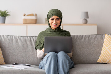 Cheery young Arab woman in hijab sits on couch in a comfortable home office. She uses her laptop for an online business webinar, confidently participating in the discussion.