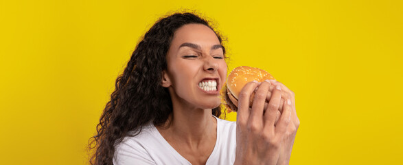 A young woman with curly hair grins widely as she holds a burger close to her face. The vibrant yellow background adds energy to the moment, showcasing her enthusiasm for the meal ahead.