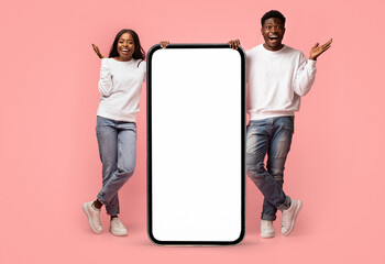 A millennial African American man and woman stand in a vibrant pink setting, showcasing a large blank cellphone. They express amazement, gesturing enthusiastically while looking at the camera.