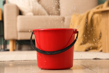 Water leaking from ceiling into red bucket in living room, closeup. Flood concept © Pixel-Shot