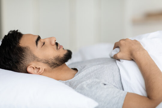 Young Arab man lies comfortably in bed with closed eyes, enjoying a deep sleep at home. The relaxing atmosphere creates a serene environment for rest and rejuvenation.