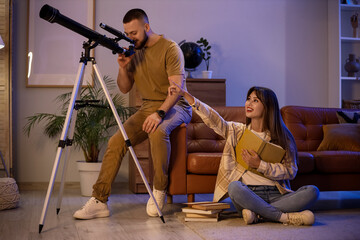 Fototapeta premium Young couple with books looking through telescope in room at night
